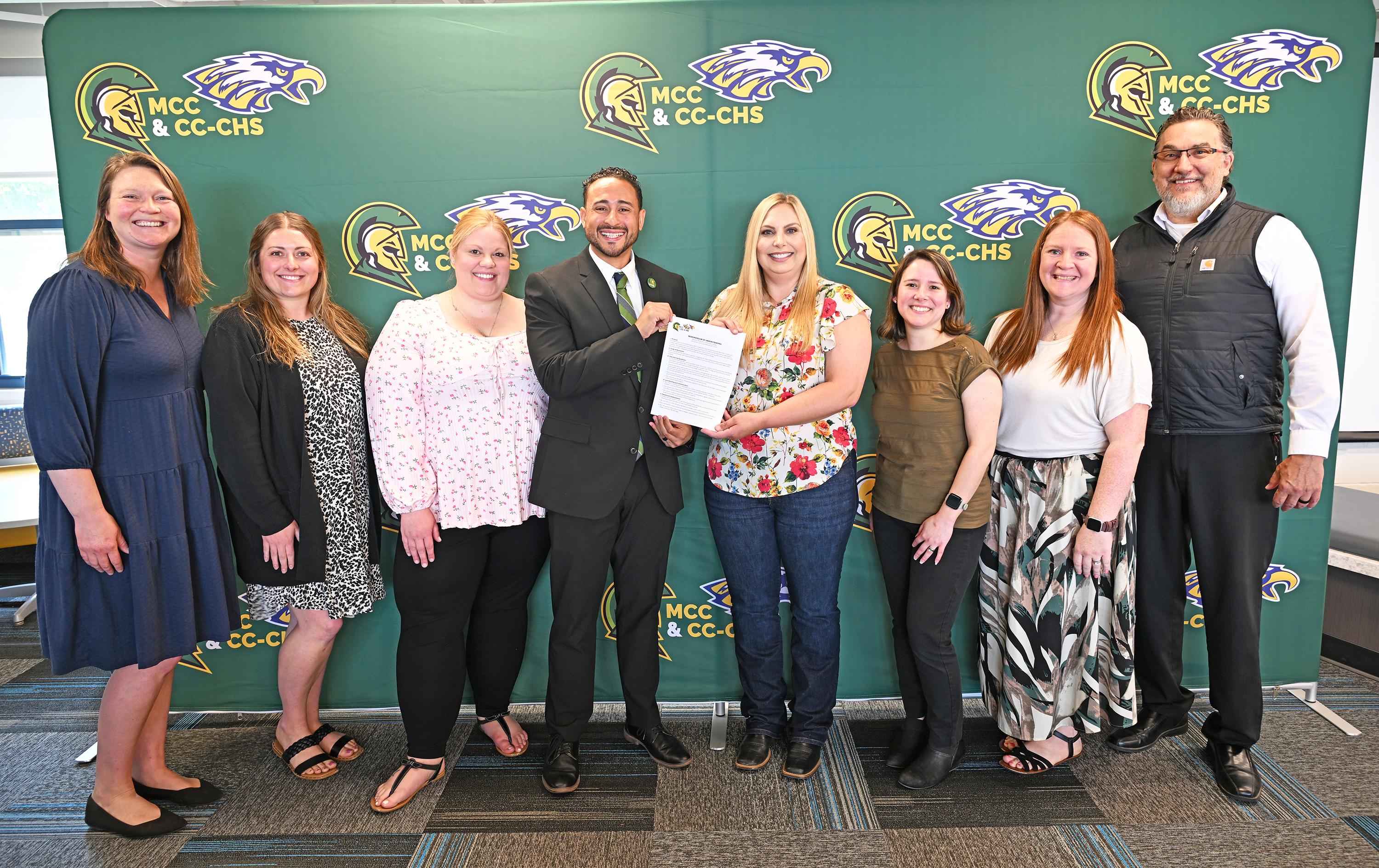 MCC staff and Carson City-Crystal schools staff pose in front of a backdrop featuring both schools logos with a paper copy of the new direct admissions agreement.
